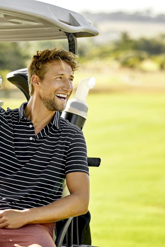 A person is sitting in a golf cart, smiling, with golf clubs in the back, on a sunny day at a golf course.
