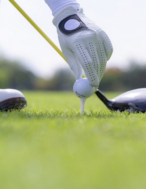 A person wearing a golf glove is placing a golf ball on a tee, with a golf club nearby on a grassy course.