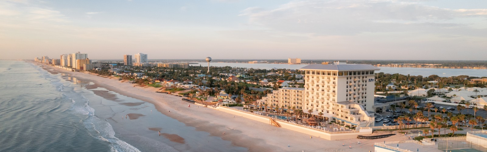 The image shows a coastal cityscape with a sandy beach, several buildings, and a calm ocean under a clear sky.