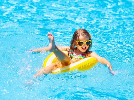 A girl wearing sunglasses and a colorful swimsuit is joyfully relaxing in a yellow float in a bright blue swimming pool.