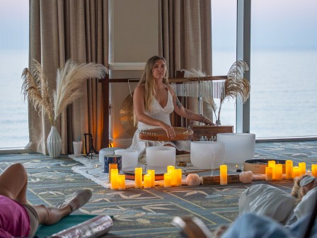 A person is sitting on the floor with sound bowls, surrounded by candles and a serene setting near a window with a sea view.