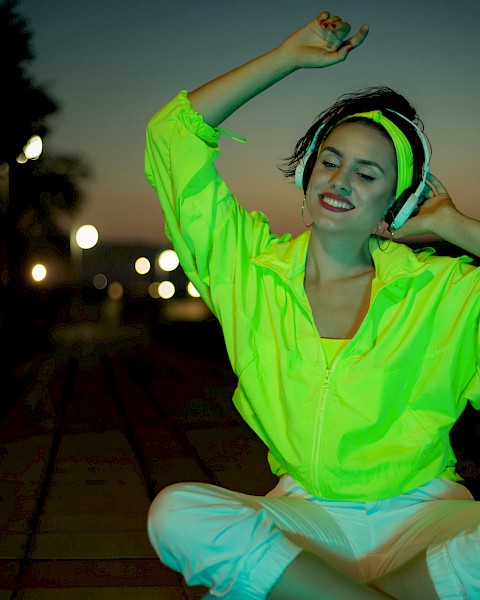 A person in a neon green jacket and white pants sits on a pathway at dusk, smiling with eyes closed, against a background of streetlights.