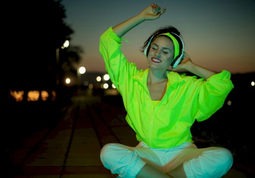 A person in a neon green jacket and white pants sits on a pathway at dusk, smiling with eyes closed, against a background of streetlights.