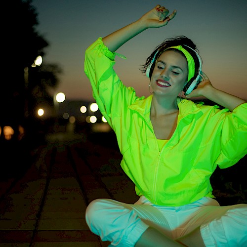A person in a neon green jacket and white pants sits on a pathway at dusk, smiling with eyes closed, against a background of streetlights.