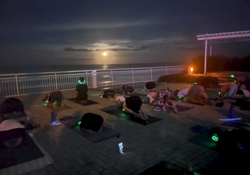 A group practicing yoga on a patio at night, with the moon illuminating the sky over a body of water in the background.