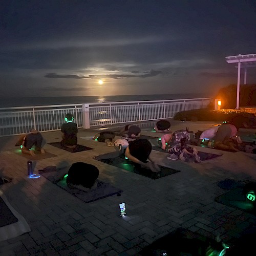 A group practicing yoga on a patio at night, with the moon illuminating the sky over a body of water in the background.
