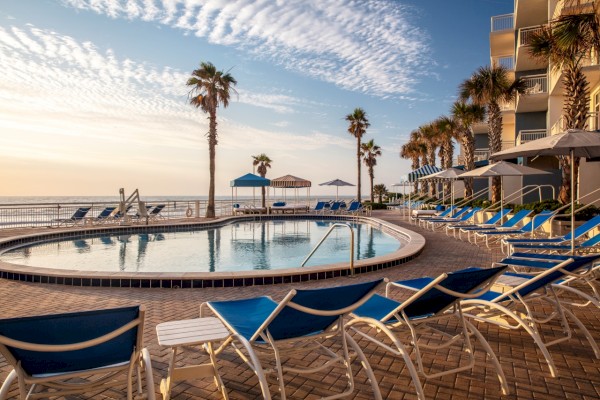 The image shows a beachfront pool area with blue lounge chairs, palm trees, and umbrellas, overlooking the sea under a clear sky.