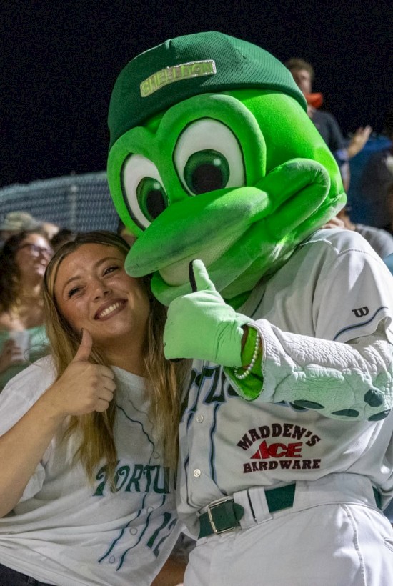 A person poses with a green frog mascot, both showing a thumbs-up, in a lively crowd setting.
