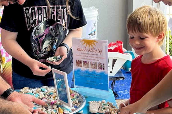 People are selecting seashells and small items from trays at a market stall. A child smiles while adults browse.