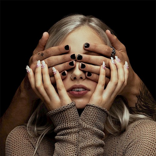 A person with platinum blonde hair is being playfully covered at the eyes by hands with black nail polish in a close-up portrait.