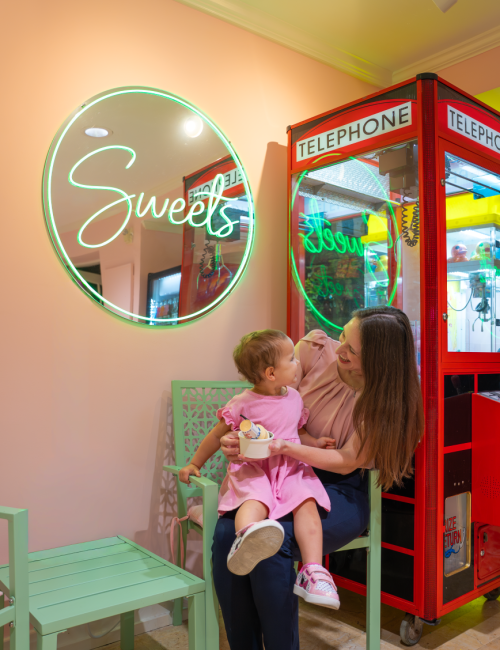 A woman and a child share a joyful moment inside a colorful sweets shop with neon signs and a red claw machine behind them.