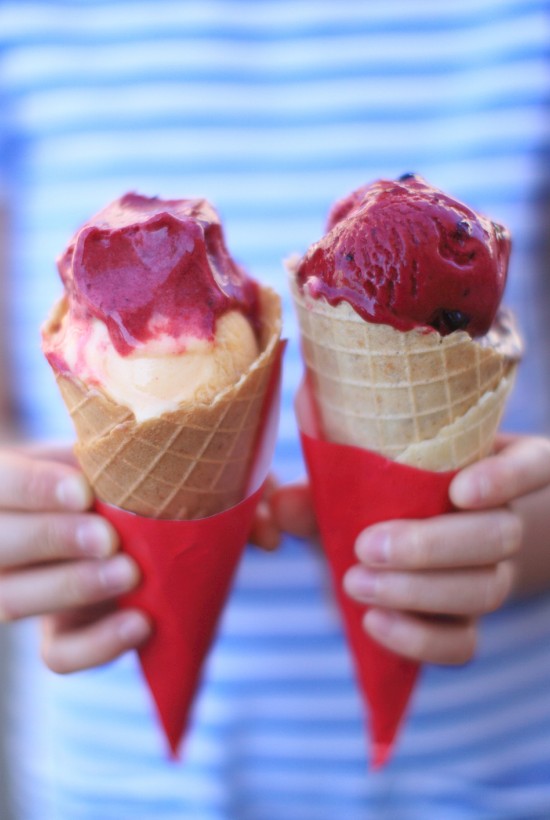 A person in a striped shirt holds two ice cream cones with red and pink scoops, wrapped in red paper, against a blurred background.
