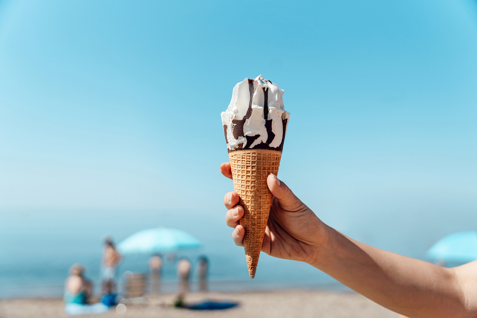 A hand holds an ice cream cone with vanilla and chocolate ice cream against a beach background with umbrellas and people relaxing by the sea.