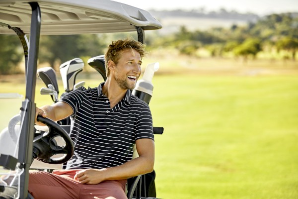 A person smiling, sitting in a golf cart on a golf course with clubs in the back.