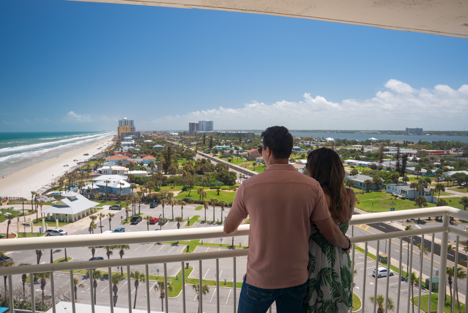 A couple is enjoying the view from a balcony overlooking a sunny beach and a coastal town with high-rise buildings.