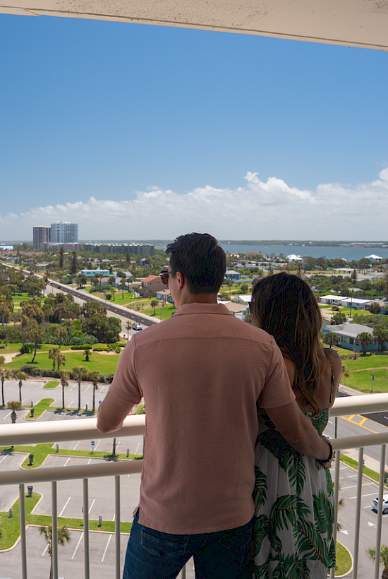 A couple stands on a balcony overlooking a beach and a coastal cityscape under a bright, partly cloudy sky.