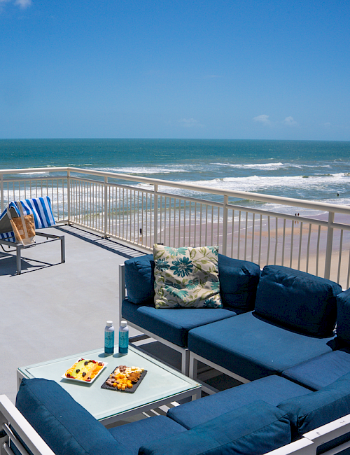 A spacious balcony with blue seating, sun loungers, and a table with snacks, overlooking a beach and the ocean under clear skies.