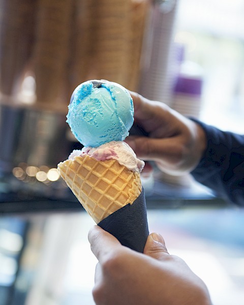 A person holds a waffle cone with blue and pink ice cream scoops in a shop setting.