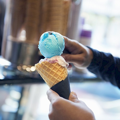 A person holds a waffle cone with blue and pink ice cream scoops in a shop setting.
