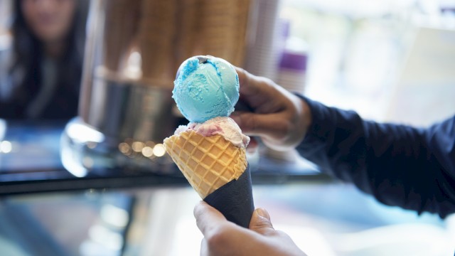 A person holds a waffle cone with blue and pink ice cream scoops in a shop setting.