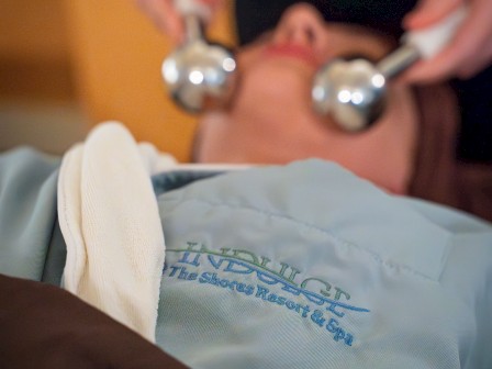 A person is receiving a facial treatment using two metal tools. The individual is wearing a blue garment with embroidered text.