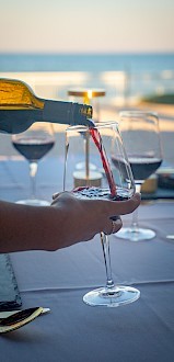 Someone is pouring red wine into a glass on a table set for dining, with a beach view in the background.