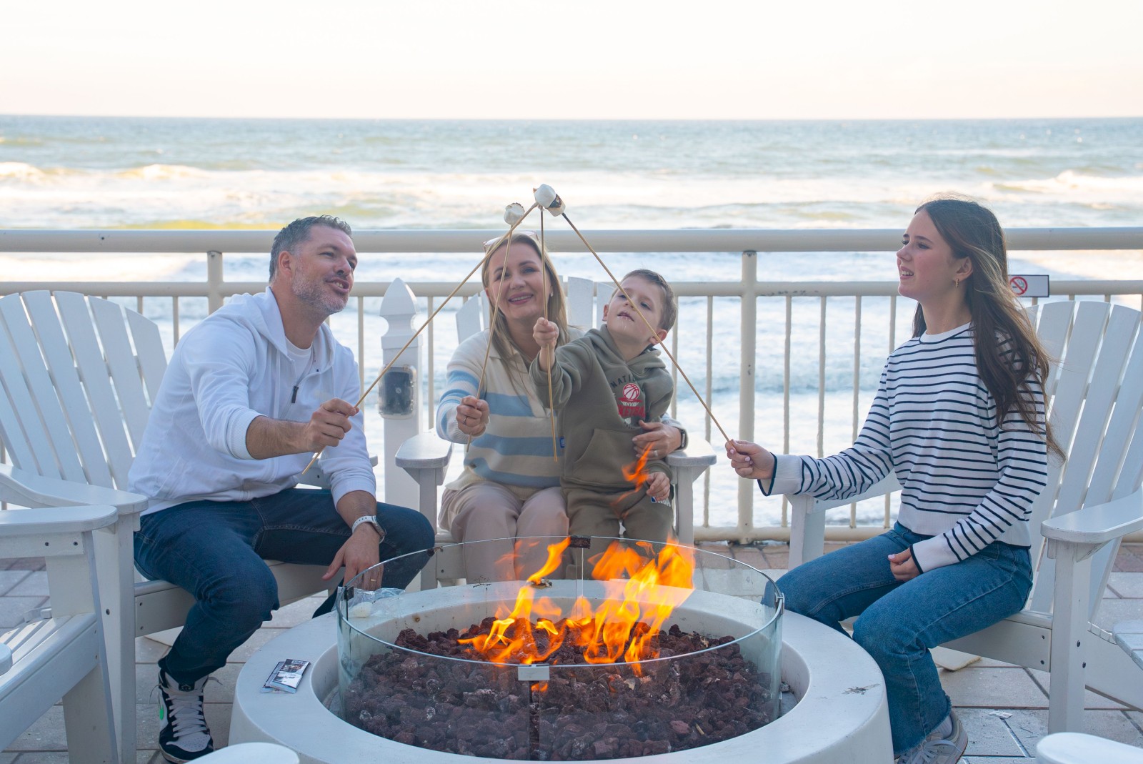 A group of people is toasting marshmallows around a fire pit by the ocean, sitting on white Adirondack chairs.
