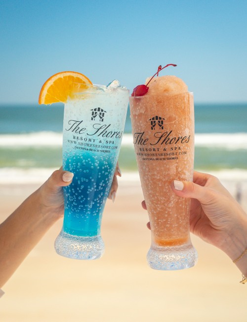 Two hands are holding colorful drinks in tall glasses at the beach, one blue with an orange slice and one pink with a cherry on top.