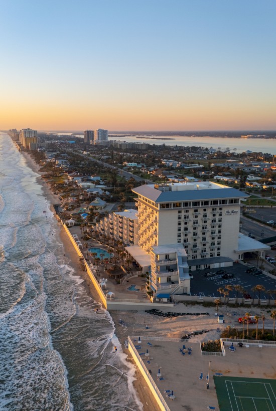 An aerial view of a coastal city at sunset showing buildings, a beach, ocean waves, and a tennis court along the shore.