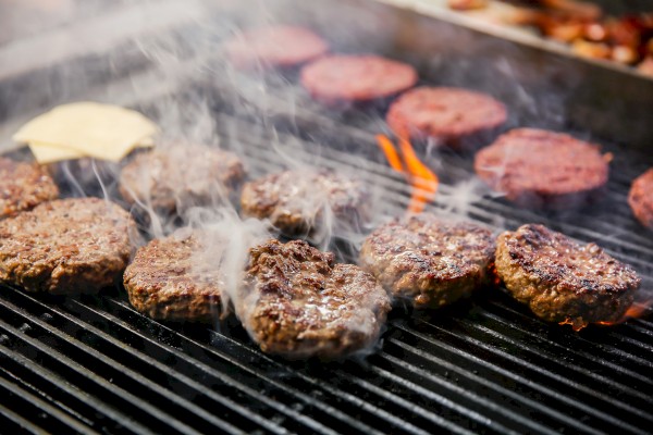 Burger patties are grilling on a barbecue, with some flames visible and smoke rising from the meat.
