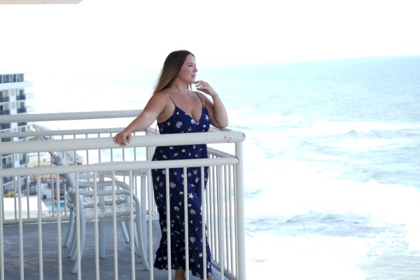A person stands on a balcony overlooking a beach with ocean views, enjoying the sunny day and coastal scenery.