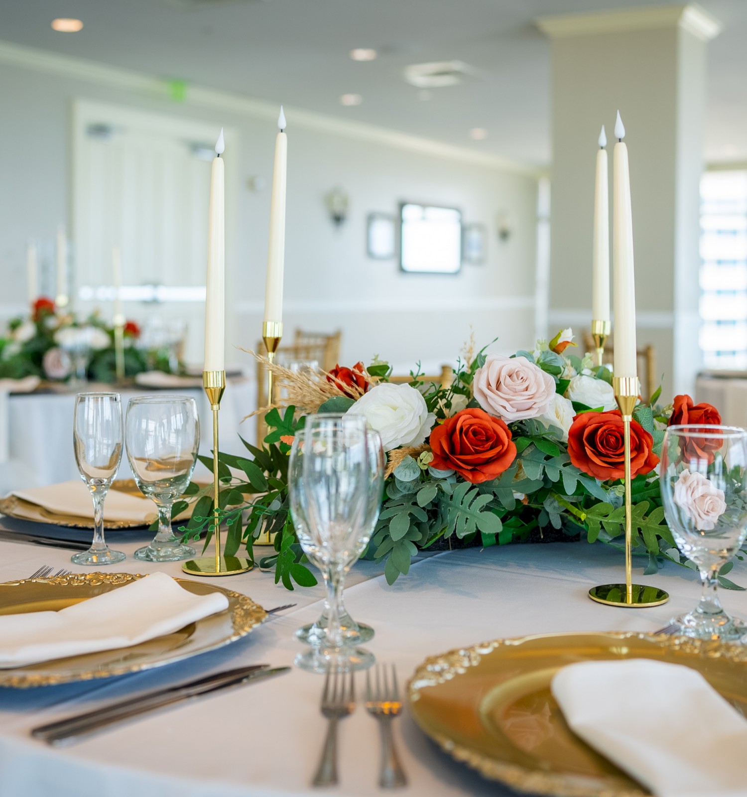 A beautifully set dining table with elegant plates, glassware, and a floral centerpiece featuring red and white roses and lit candles.