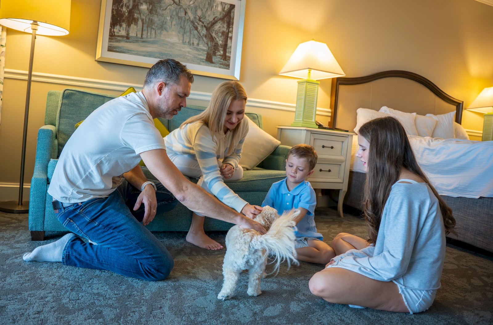 A family of four is sitting on the floor in a cozy room, smiling and petting a small dog, while soft lighting creates a warm atmosphere.
