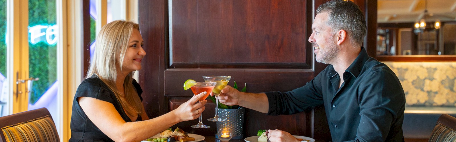 A couple is dining in a restaurant, toasting with drinks over a table. The setting is cozy with wooden decor.