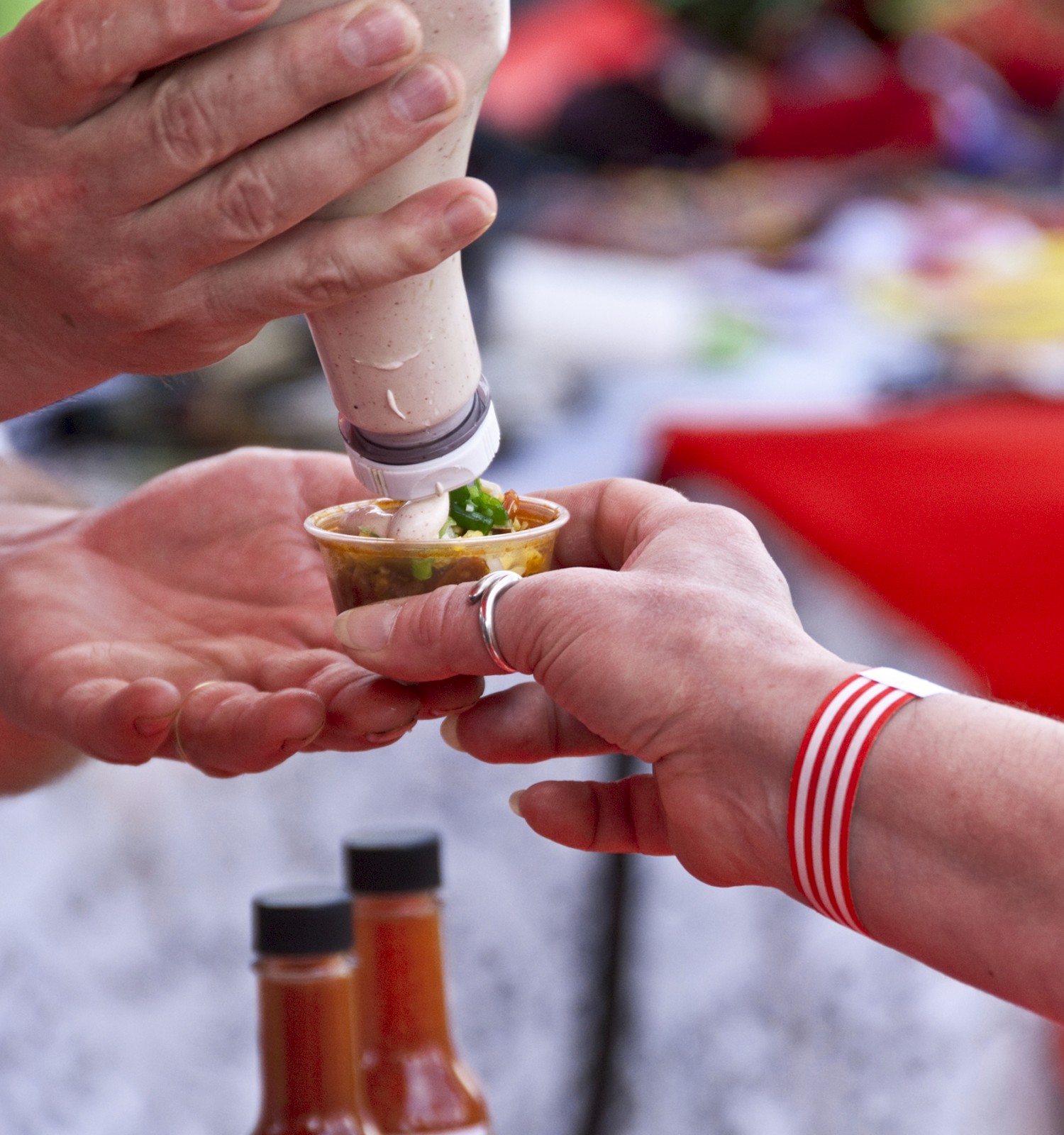 A person pours white sauce into a small cup held by another person, with hot sauce bottles visible in the background.