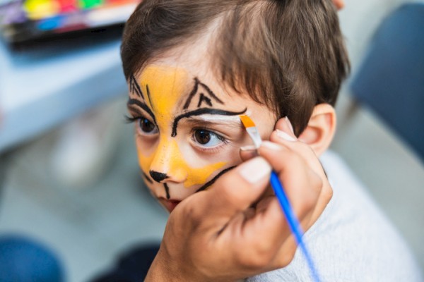 A child is getting their face painted as a cute animal, likely a cat or tiger, by an artist with a small brush.