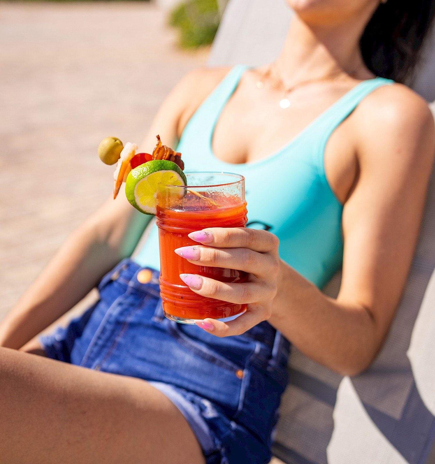 A person in sunglasses relaxes outdoors, holding a garnished red drink. They wear a light blue top and denim shorts.