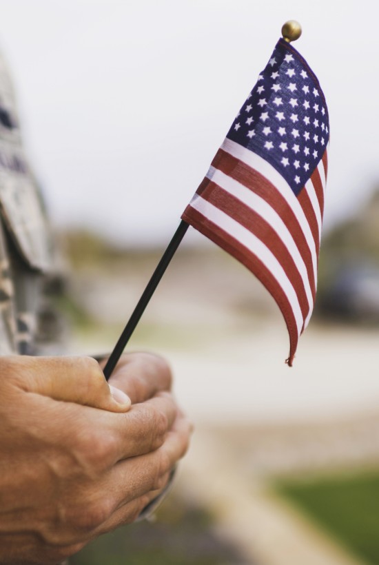Close-up of a person in military uniform holding a small American flag outdoors.