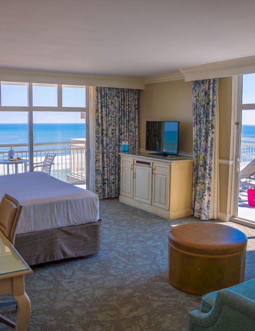 Oceanfront hotel room with a king bed, desk, TV, sofa, and large windows opening to a balcony with views of the Atlantic Ocean.