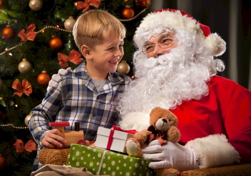 A young boy smiles while sitting beside Santa Claus, who is holding a teddy bear and wrapped presents. A decorated Christmas tree with warm lights and ornaments glows softly in the background, capturing a joyful holiday moment.
