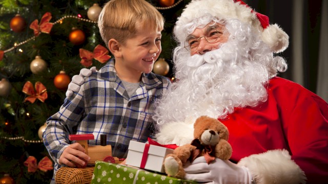 A young boy smiles while sitting beside Santa Claus, who is holding a teddy bear and wrapped presents. A decorated Christmas tree with warm lights and ornaments glows softly in the background, capturing a joyful holiday moment.