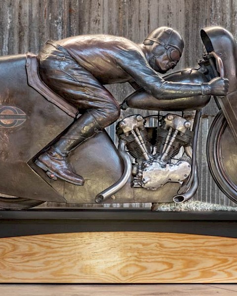 Bronze sculpture of legendary motorcycle racer Joe Petrali riding his streamlined Harley-Davidson EL, displayed on a wooden and metal base in front of a rustic corrugated metal wall.