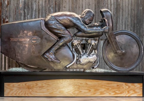 Bronze sculpture of legendary motorcycle racer Joe Petrali riding his streamlined Harley-Davidson EL, displayed on a wooden and metal base in front of a rustic corrugated metal wall.