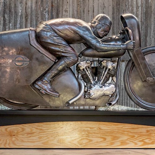 Bronze sculpture of legendary motorcycle racer Joe Petrali riding his streamlined Harley-Davidson EL, displayed on a wooden and metal base in front of a rustic corrugated metal wall.