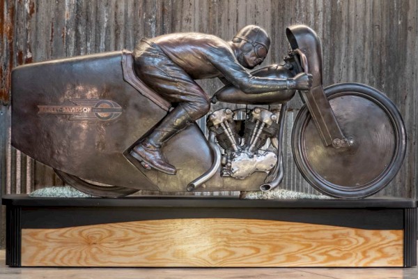 Bronze sculpture of legendary motorcycle racer Joe Petrali riding his streamlined Harley-Davidson EL, displayed on a wooden and metal base in front of a rustic corrugated metal wall.