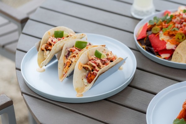 Three tacos with grilled meat, shredded lettuce, cheese, and lime wedges on a white plate, outdoors on a wooden table.