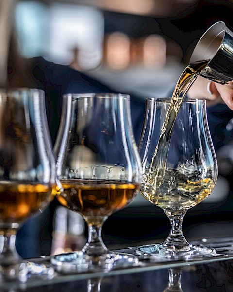 A person is pouring amber liquid from a jigger into a glass, alongside several similar glasses on a bar counter.