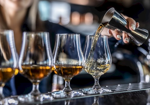 A person is pouring amber liquid from a jigger into a glass, alongside several similar glasses on a bar counter.