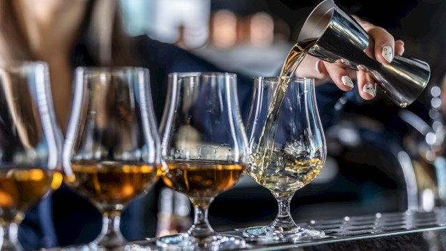 A person is pouring amber liquid from a jigger into a glass, alongside several similar glasses on a bar counter.