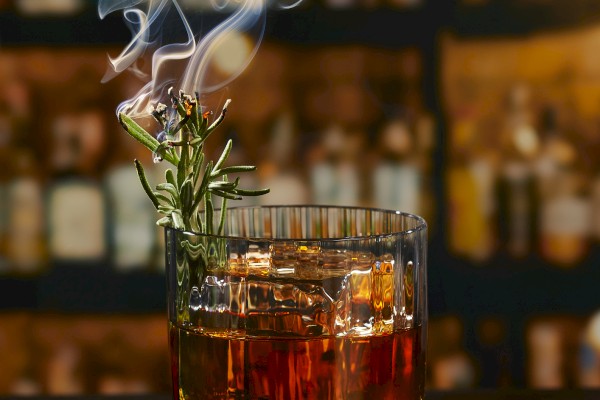 A cocktail in a glass with smoke, garnished with rosemary and an orange slice, placed on a dark surface against a blurred background.
